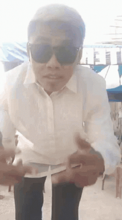 A Man Wearing Sunglasses And A White Shirt Is Standing On A Sidewalk . GIF