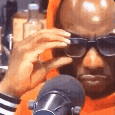 A Man Wearing Sunglasses And An Orange Hoodie Is Sitting In Front Of A Microphone . GIF