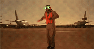 A Man Wearing Sunglasses And An Orange Vest Stands In Front Of Planes GIF