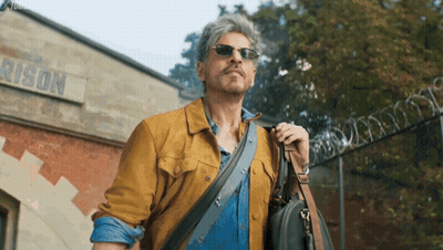 A Man Wearing Sunglasses And Carrying A Briefcase Walks In Front Of A Building That Says Prison GIF