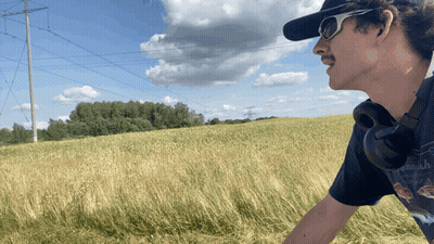 A Man Wearing Sunglasses And Headphones Stands In A Field Of Tall Grass GIF