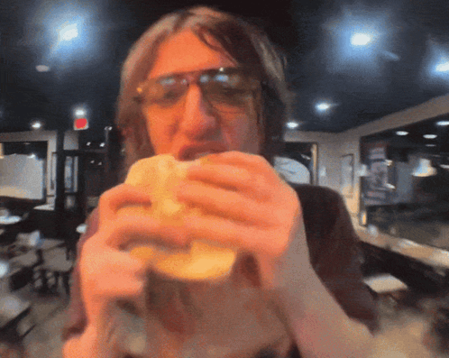 A Man Wearing Sunglasses Eating A Sandwich In A Restaurant GIF