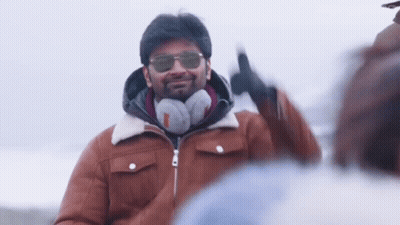 A Man Wearing Sunglasses , Headphones And A Brown Jacket Is Smiling And Giving A Peace Sign . GIF