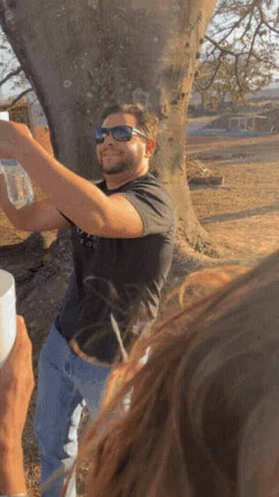 A Man Wearing Sunglasses Holds A Bottle Of Water In Front Of A Tree GIF