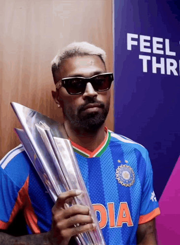 A Man Wearing Sunglasses Holds A Trophy In Front Of A Wall That Says Feel Thr GIF