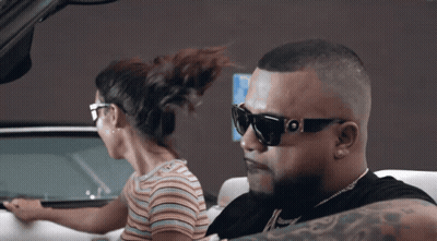 A Man Wearing Sunglasses Is Driving A Car Next To A Woman In A Striped Shirt GIF
