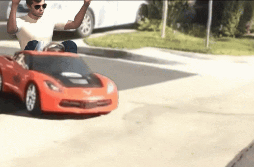 A Man Wearing Sunglasses Is Driving A Red Toy Car GIF