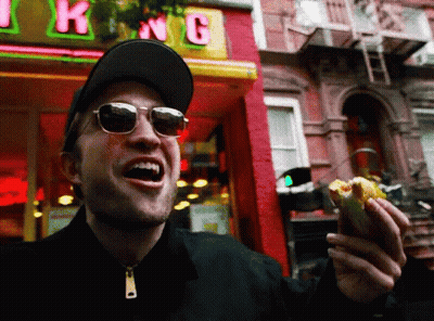 A Man Wearing Sunglasses Is Eating A Hot Dog In Front Of A Store Called King GIF