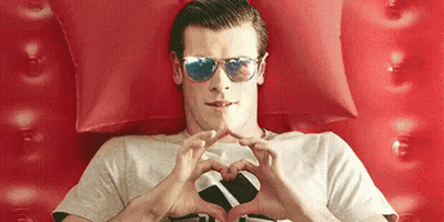 A Man Wearing Sunglasses Is Making A Heart Shape With His Hands While Laying On A Bed . GIF