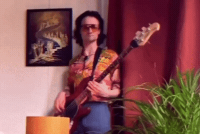 A Man Wearing Sunglasses Is Playing A Bass Guitar In A Living Room . GIF