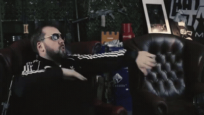 A Man Wearing Sunglasses Is Sitting In A Chair In Front Of A Sign That Says Am GIF