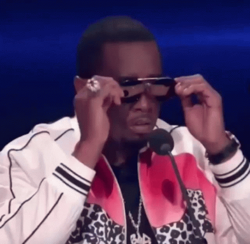 A Man Wearing Sunglasses Is Sitting In Front Of A Microphone And Adjusting His Sunglasses . GIF