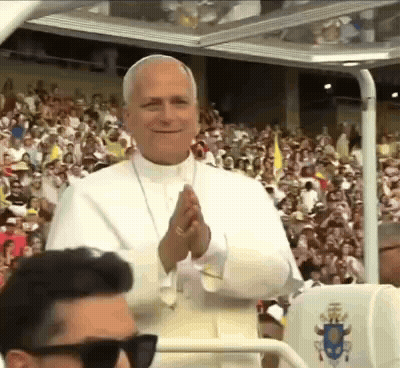 A Man Wearing Sunglasses Is Standing In Front Of A Crowd Of People Applauding The Pope . GIF