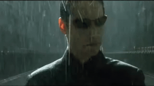 A Man Wearing Sunglasses Is Standing In The Rain Looking At The Camera . GIF