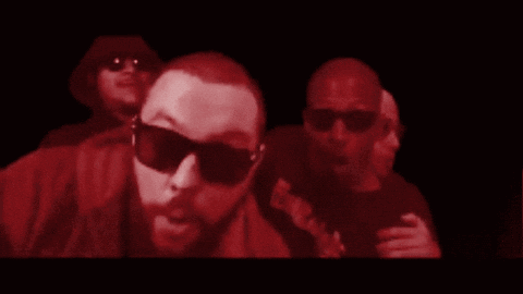 A Man Wearing Sunglasses Is Surrounded By Other Men In A Dark Room GIF