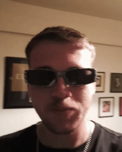 A Man Wearing Sunglasses Looks At The Camera In Front Of A Wall With Pictures On It GIF