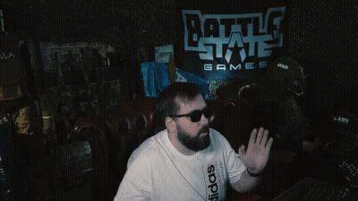 A Man Wearing Sunglasses Sits In Front Of A Battle State Games Sign GIF