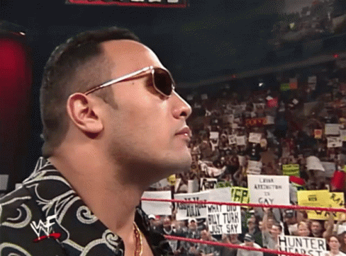 A Man Wearing Sunglasses Stands In A Wrestling Ring With A Crowd Behind Him GIF