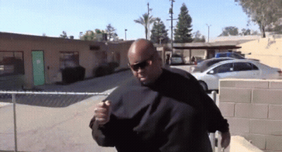 A Man Wearing Sunglasses Stands In Front Of A Building With A Green Door And Cars Parked In Front Of It GIF