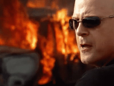 A Man Wearing Sunglasses Stands In Front Of A Burning Building GIF