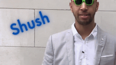 A Man Wearing Sunglasses Stands In Front Of A Wall That Has The Word Shush Painted On It GIF