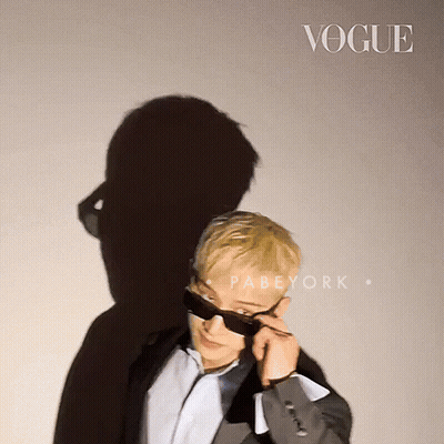 A Man Wearing Sunglasses Stands In Front Of A Wall That Says Vogue GIF