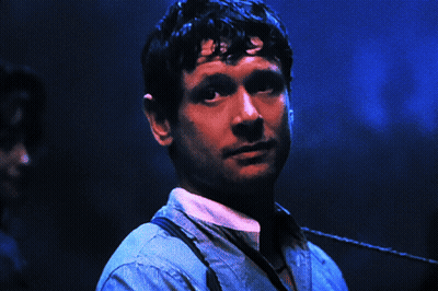 A Man Wearing Suspenders Looks At The Camera With A Blue Background GIF