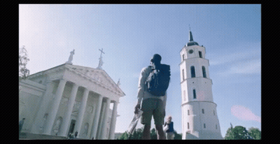 A Man With A Backpack Stands In Front Of A Large Building With Columns And A Clock Tower GIF