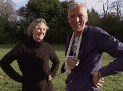 A Man With A Bandage On His Arm Stands Next To A Woman In A Black Sweater GIF