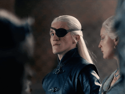 A Man With A Bandage On His Eye Stands Next To A Woman GIF