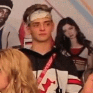 A Man With A Bandage On His Head Is Standing In Front Of A Group Of People . GIF