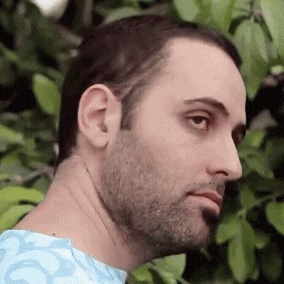 A Man With A Beard And A Blue Shirt Is Standing In Front Of A Tree And Looking At The Camera . GIF