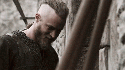 A Man With A Beard And A Chain Around His Neck Is Leaning Against A Wall GIF