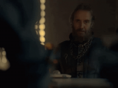 A Man With A Beard And A Chain Around His Neck Is Sitting At A Table With Other People . GIF