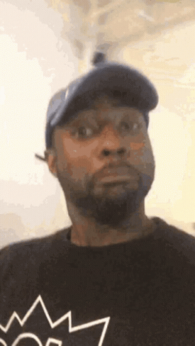 A Man With A Beard And A Hat Is Wearing A Black Shirt With A Crown On It . GIF