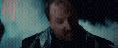 A Man With A Beard And A Leather Jacket Is Standing In A Dark Room With Smoke Coming Out Of His Mouth . GIF