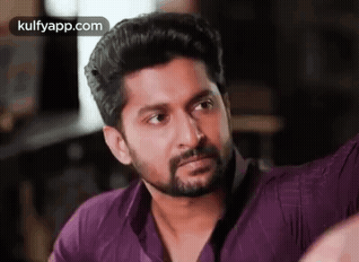 A Man With A Beard And A Purple Shirt Is Looking At The Camera . GIF