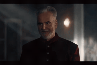 A Man With A Beard And A Red Jacket Is Standing In A Dark Room And Smiling . GIF