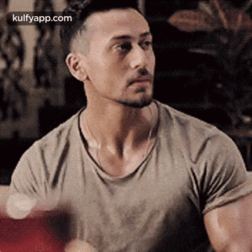 A Man With A Beard And A T-Shirt Is Sitting At A Table And Looking At The Camera . GIF