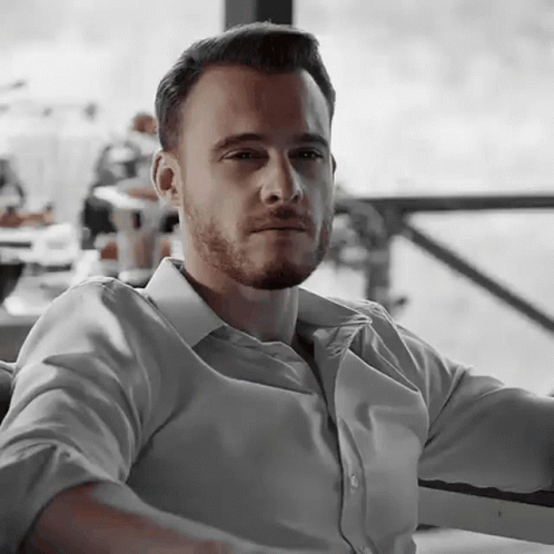 A Man With A Beard And A White Shirt Is Sitting In A Chair . GIF
