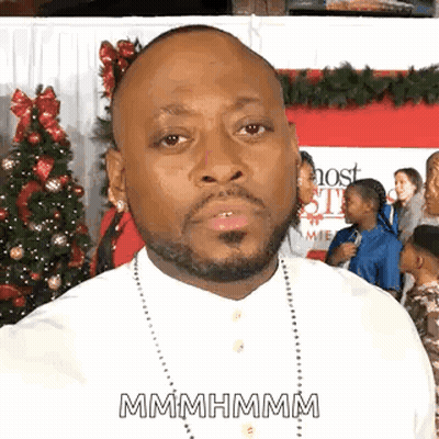 A Man With A Beard And A White Shirt Is Standing In Front Of A Christmas Tree . GIF