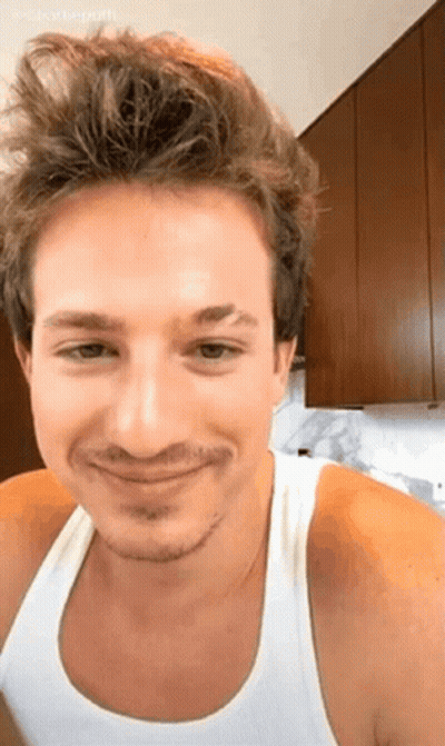 A Man With A Beard And A White Tank Top Is Smiling In A Kitchen . GIF