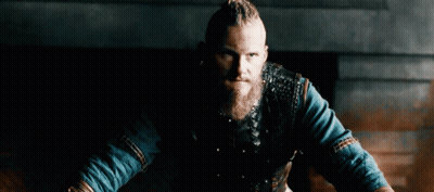 A Man With A Beard And Braided Hair Is Wearing A Leather Vest And A Blue Shirt . GIF