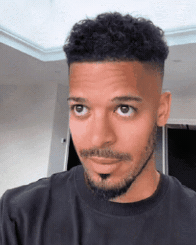 A Man With A Beard And Curly Hair Is Wearing A Black Shirt And Looking At The Camera . GIF