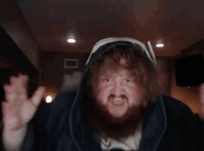 A Man With A Beard And Curly Hair Is Wearing A Hooded Jacket And Making A Funny Face . GIF