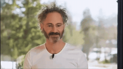 A Man With A Beard And Curly Hair Is Wearing A White T-Shirt And A Microphone . GIF