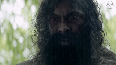 A Man With A Beard And Curly Hair Looks At The Camera With A Logo For A Company In The Background GIF