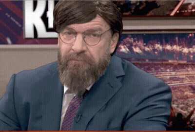 A Man With A Beard And Glasses Is Sitting In Front Of A Wall With The Letter K On It GIF