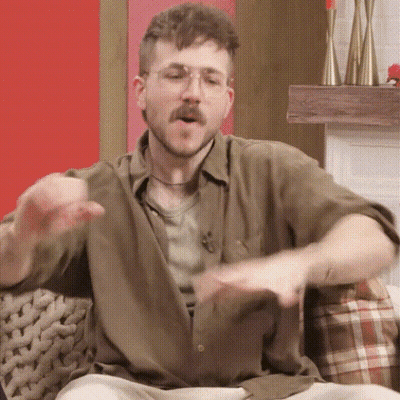 A Man With A Beard And Glasses Is Sitting On A Couch And Making A Funny Face . GIF