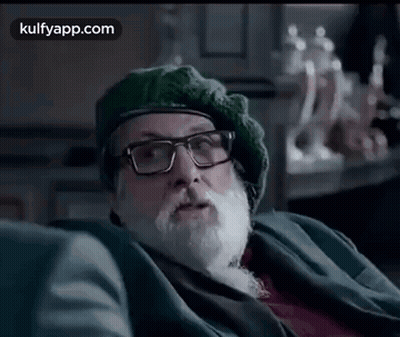 A Man With A Beard And Glasses Is Sitting On A Couch Wearing A Green Hat . GIF